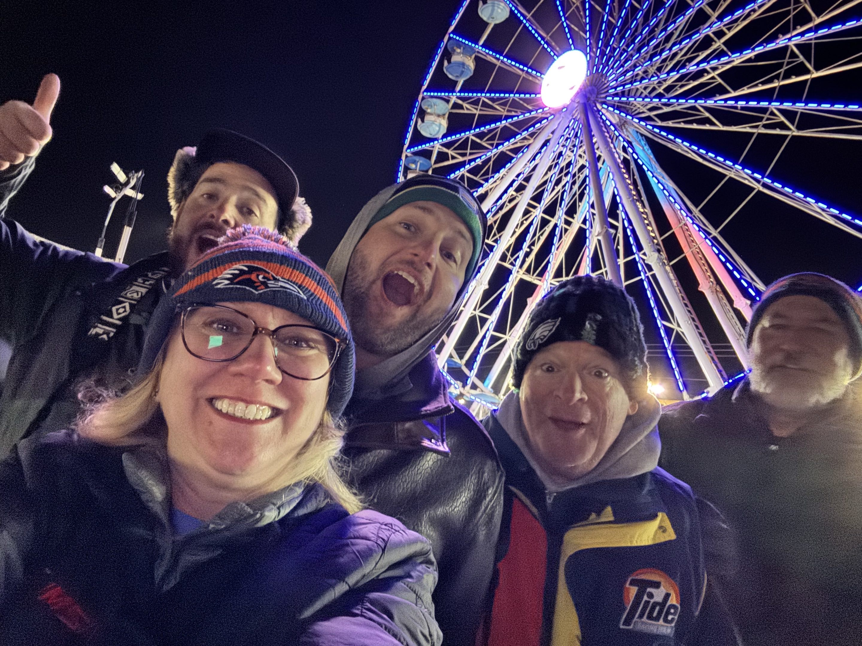 Cold people taking a selfie in front of a neon-lit ferris wheel at night